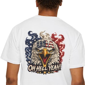 Può includere: Una t-shirt bianca a maniche corte con una grafica sul retro raffigurante una testa di aquila calva con il becco giallo. L'aquila è circondata da motivi di fumo rosso, bianco e blu. Il testo USA è sulla fronte e OH HELL YEAH! è in basso.