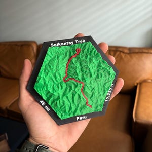 May include: A hexagonal 3D printed topographic map of the Salkantay Trek in Peru. The terrain is bright green with a raised red line indicating the trail. The black border features white text reading Salkantay Trek, 46 mi, 15,255 ft, and Peru.