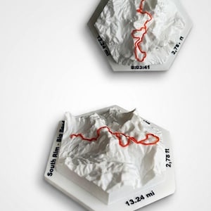 May include: Two white hexagonal 3D relief maps of the South Rim in Big Bend. Each map features a raised orange trail line, with black text displaying South Rim - Big Bend, 13.24 mi, 2,787 ft, and 8:03:41 printed on the flat white edges of the base.