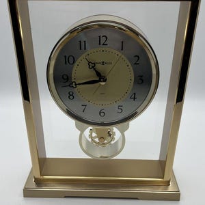 May include: A rectangular gold-tone metal mantel clock with a clear glass frame. The circular clock face features a silver outer ring with black numerals, a gold center, and black hands. The text Howard Miller is printed on the face.
