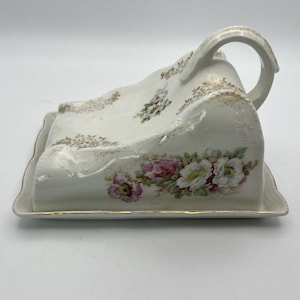 May include: A white ceramic butter dish with a domed cover featuring a handle. The piece is decorated with a floral pattern of pink and white flowers on the side and gold leaf accents along the edges and top surface of the domed cover.