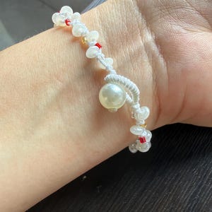 May include: A white cord bracelet featuring a series of small white beads, interspersed with tiny red and gold accent beads, and a single larger, prominent white pearl-like bead attached to a looped section of the white cord.