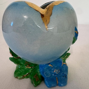 May include: A heart-shaped ceramic vase with a light blue crackle glaze finish. The top edge features a gold-painted accent. The base is decorated with sculpted green leaves and a bright blue flower, both detailed with gold trim along the edges.
