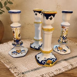 Puede incluir: Cuatro portavelas de cerámica blanca con motivos florales y geométricos pintados a mano en azul, amarillo y verde. Cada candelabro presenta una base ensanchada, una columna central delgada con relieves decorativos y un borde superior ancho.