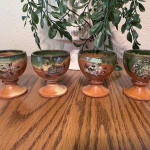 Può includere: Quattro portauova in ceramica con base a piedistallo, caratterizzati da un design bicolore con fondo color terracotta e bordo verde scuro. Ogni tazza presenta una stampa grafica circolare unica e dettagliata di vari luoghi e scene.