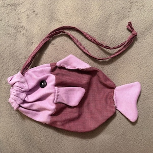 May include: A fish-shaped fabric pouch featuring a light purple head and tail with a dark maroon checkered body. The pouch includes a black button eye, a drawstring closure at the mouth, and a long, thin matching maroon fabric strap for carrying.