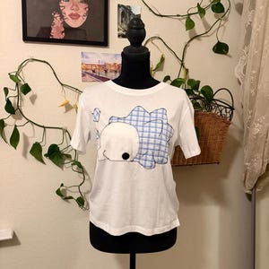 May include: A white short-sleeved t-shirt featuring a large, central fabric appliqué of a stylised creature. The creature has a solid white body with a black circular eye and a blue and white checkered pattern on its back, all outlined in dark stitching.