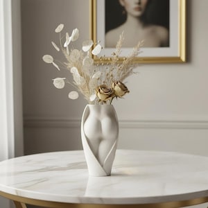 May include: A white ceramic vase shaped like a human torso, featuring a smooth, matte finish. The vase is filled with a dried floral arrangement consisting of beige pampas grass, round silver dollar eucalyptus leaves, and two light brown roses.