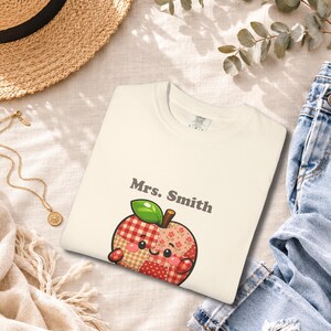 Może przedstawiać: Kremowy t-shirt z grafiką uśmiechniętego jabłka z patchworkowym wzorem w odcieniach czerwieni, różu i beżu. Jabłko ma zielony listek, a nad ilustracją jabłka nadrukowany jest ciemnoszary napis "Mrs. Smith".