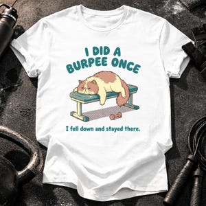 May include: A white short-sleeve t-shirt featuring a graphic of a fluffy brown and cream cat lying on a gym bench. The text reads I DID A BURPEE ONCE and I fell down and stayed there. A small brown dumbbell is illustrated on the floor below the bench.
