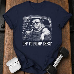May include: A navy blue short-sleeve t-shirt featuring a white graphic illustration of a bulldog sitting in a car with its arm resting on the door. Below the illustration, the text reads OFF TO PUMP CHEST in a bold, white, capitalized font.