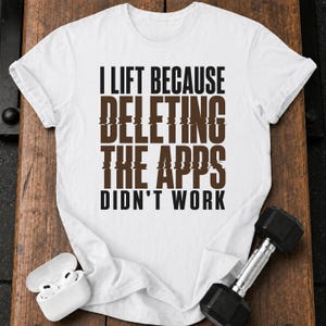 May include: A white short-sleeved t-shirt featuring the black and brown text I LIFT BECAUSE DELETING THE APPS DIDN'T WORK. The shirt is displayed alongside a pair of white wireless earbuds in a charging case and a black metal dumbbell.