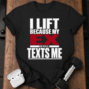 May include: A black short-sleeved t-shirt featuring white and red text that reads I LIFT BECAUSE MY EX STILL TEXTS ME. The shirt is displayed alongside a pair of white wireless earbuds in a charging case and a single black metal dumbbell.