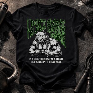 May include: A black t-shirt featuring a graphic of a muscular dog wearing a spiked collar and holding chains. The text reads, "I DON'T SWEAT, I'M JUST LEAKING DESPAIR" in green and "MY DOG THINKS I'M A HERO, LET'S KEEP IT THAT WAY." in white.