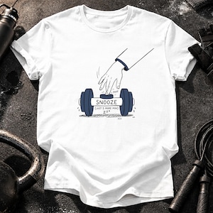 May include: A white short-sleeved t-shirt featuring a graphic illustration of a hand pressing a button on a dumbbell. The button is labeled SNOOZE, with the text (JUST 5 MORE MIN) and ZZZ below it, and the word POOF near the finger.