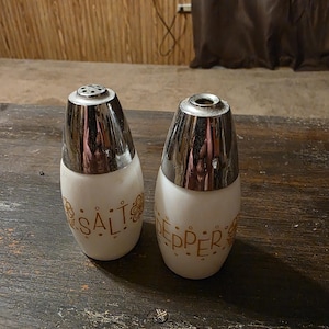 May include: A pair of salt and pepper shakers featuring white glass bodies with gold floral patterns and the words SALT and PEPPER printed in gold. Each shaker is topped with a polished chrome metal cap, with the salt shaker having multiple holes.