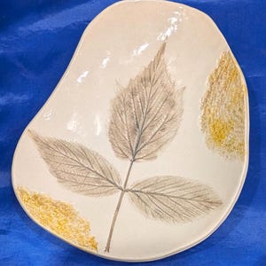 May include: An off-white ceramic dish with an irregular, organic shape. The surface features a detailed, pressed botanical impression of a three-part leaf in muted gray tones, accented by textured patches of yellow glaze on the edges.