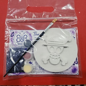 May include: A craft kit in a clear plastic bag containing a round white plaster disc with a raised skull and crossbones design, a black-handled paintbrush, three small circular paint pots in black, pink, and yellow, and a small bag of dark glitter.