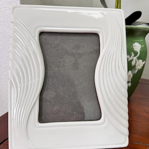 Può includere: Una cornice portafoto rettangolare in ceramica bianca con finitura lucida e motivi ondulati in rilievo lungo i bordi verticali sinistro e destro. L'apertura centrale è rettangolare con lati leggermente curvi, che incornicia un inserto grigio scuro.