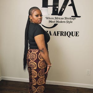 Puede incluir: Una camiseta negra de manga corta combinada con pantalones anchos con un patrón de espirales circulares en marrón oscuro y tostado. La pared detrás muestra el texto HA Where African Heritage Meet Modern Style y ZA AFRIQUE en negro.