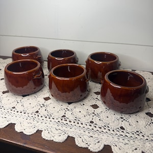 May include: A set of six small, round, brown glazed ceramic bowls. Each bowl features a smooth, glossy finish with a deep chocolate brown color and small, integrated side handles. The bowls are arranged in a group on a white surface.