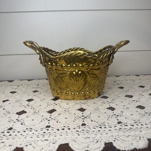 May include: A polished brass oval basket featuring a raised shell motif on the front, a beaded border along the base, and a twisted rope-style rim. The vessel includes two ornate handles on the sides, showcasing a metallic gold finish throughout.