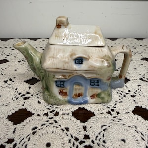 May include: A ceramic teapot shaped like a cottage with a removable roof lid. The house features a blue arched doorway, small dark windows, a green textured base, and a tan roof. A curved handle is on the right and a spout extends from the left.