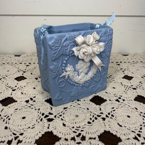 May include: A square light blue ceramic planter featuring a raised white cameo of a profile figure surrounded by embossed ivy leaves. The planter is adorned with a white sculpted rose cluster and ribbon detail, with light blue ribbons attached to the top corners.