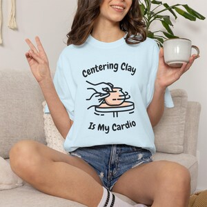 Peut inclure: Un t-shirt bleu clair avec une illustration de mains façonnant un pot en argile sur un tour de potier. Le texte au-dessus indique Centering Clay et le texte en dessous indique Is My Cardio. Le motif est en noir, pêche et blanc.
