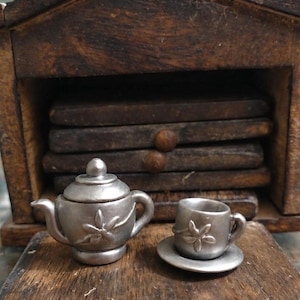 Könnte beinhalten: Eine Miniatur-Teekanne aus silberfarbenem Metall mit Deckel und eine passende Teetasse auf einer Untertasse. Beide Teile haben ein erhabenes Blumenmuster auf der Vorderseite. Die Teekanne hat einen geschwungenen Ausguss und Griff, die Tasse steht auf einem kleinen Teller.