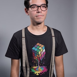 May include: A black short-sleeved t-shirt featuring a graphic of a colorful cube puzzle that appears to be melting into a vibrant puddle of paint. The shirt is worn with beige suspenders and black-rimmed glasses are worn on the face.