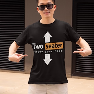 Può includere: Una t-shirt nera a maniche corte con una freccia bianca rivolta verso l'alto sopra la parola Two, un rettangolo giallo contenente la parola seater e una freccia bianca rivolta verso il basso sotto il testo enjoy your ride, stampati sul davanti.