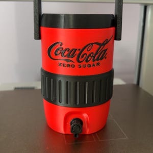 Op de afbeelding: Een felrode drankkoeler met een zwarte handgreep, een zwart deksel en een zwarte, getextureerde gripband in het midden. De voorzijde toont het zwarte Coca-Cola-logo met de tekst Zero Sugar eronder en een kleine zwarte tapkraan aan de onderkant.