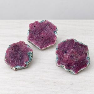 May include: Three raw ruby crystal slices with deep magenta and purple hues. Each piece features a flat, polished surface with a crystalline texture and natural, rough, greenish-grey edges that define the irregular, organic shape of the stones.