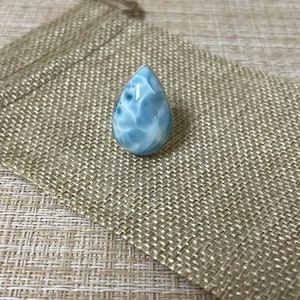 May include: A teardrop-shaped cabochon stone featuring a marbled pattern of light blue, sky blue, and white tones. The stone is set in a smooth, polished silver-colored metal frame, creating a ring with a distinct, organic, and smooth surface texture.
