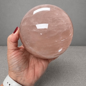 May include: A large, smooth, spherical rose quartz crystal with a translucent, pale pink hue. The surface of the stone displays natural internal veining and cloud-like inclusions, with bright white light reflections visible on the polished exterior.