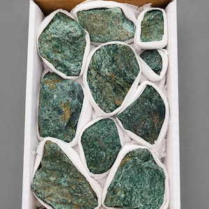 May include: A rectangular cardboard box containing nine rough, irregular green stones. Each stone is individually wrapped in white tissue paper, creating a protective barrier between the textured, dark green mineral surfaces and the box interior.