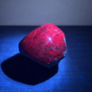May include: A polished, irregular rock specimen displaying a vibrant, glowing red surface under ultraviolet light. The stone features dark, mottled inclusions and a smooth, reflective texture across its rounded, asymmetrical form.
