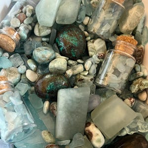 May include: A collection of light blue, green, and brown tumbled stones and polished gems mixed with three small clear glass bottles filled with tiny light blue stone chips, each bottle sealed with a light brown cylindrical cork stopper.