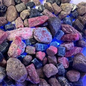 May include: A collection of raw, unpolished stones in shades of deep pink, magenta, and dark blue. The stones exhibit a rough, textured surface with some pieces showing a bright, glowing blue fluorescence under ultraviolet light exposure.