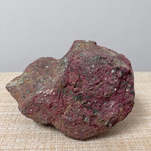 May include: A raw, unpolished mineral specimen featuring a vibrant magenta and deep red coloration with embedded dark green and metallic silver flecks throughout its rough, uneven, and textured surface, showcasing a natural crystalline structure.