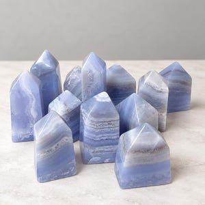 May include: A collection of twelve blue lace agate crystal towers, each featuring a pointed top and a polished finish. The stones display horizontal bands of light blue, white, and grey, with varying levels of translucency and natural mineral patterns.