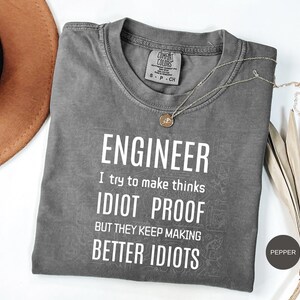 Op de afbeelding: Een antracietgrijs T-shirt met korte mouwen en witte tekst die luidt ENGINEER I try to make thinks IDIOT PROOF BUT THEY KEEP MAKING BETTER IDIOTS. Het shirt heeft een ronde hals en een Comfort Colors merklabel in de binnenkraag.