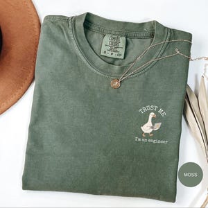 Könnte beinhalten: Ein moosgrünes Kurzarm-T-Shirt mit Rundhalsausschnitt. Auf der Brust befindet sich eine kleine Grafik einer weißen Gans, die einen Laptop hält, mit dem Text "TRUST ME I'm an engineer" in einem Bogen darüber und darunter. Das Innenetikett zeigt Comfort Colors.