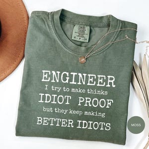 Op de afbeelding: Een mosgroen t-shirt met korte mouwen, een ronde hals en een witte grafische print met de tekst: ENGINEER I try to make thinks IDIOT PROOF but they keep making BETTER IDIOTS. Het shirt is voorzien van een Comfort Colors label in de nek.