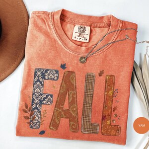 Könnte beinhalten: Ein kurzärmeliges T-Shirt in gebranntem Orange mit Rundhalsausschnitt und einem Comfort Colors Markenetikett. Auf der Vorderseite steht das Wort FALL in großen Blockbuchstaben, die jeweils mit verschiedenen Mustern wie blauem Damast, Paisley, Karo und Herbstblättern versehen sind.