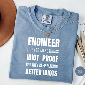 May include: A blue jean coloured short-sleeve t-shirt with white text that reads: ENGINEER I TRY TO MAKE THINKS IDIOT PROOF BUT THEY KEEP MAKING BETTER IDIOTS. The shirt features a Comfort Colors brand label at the inner neckline and a crew neck collar.