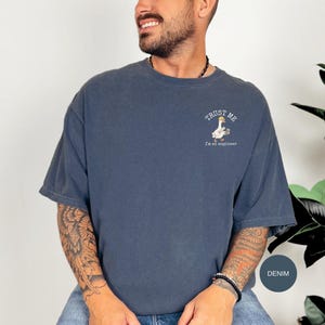 Puede incluir: Una camiseta de manga corta color denim con un pequeño gráfico en el pecho izquierdo. El gráfico muestra un ganso blanco con un casco de obra amarillo que sostiene un plano, con el texto "TRUST ME I'm an engineer" impreso en color blanco.