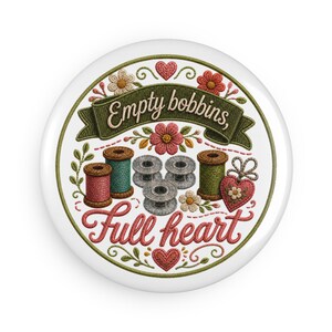 May include: A round pin featuring an embroidered design of sewing spools, bobbins, and flowers on a white background. The text reads Empty bobbins, Full heart in a cursive script. The design includes red and pink hearts and green foliage accents.