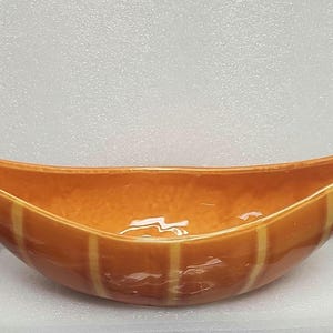 May include: A long, boat-shaped ceramic bowl with a glossy orange glaze and vertical cream-colored stripes. The vessel features elongated, pointed ends that curve upward, creating a distinct crescent silhouette with a smooth, reflective surface finish.